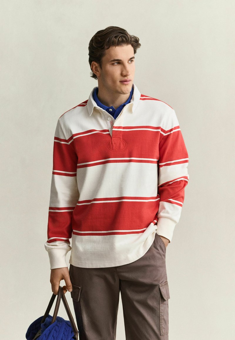 Junger Mann trägt ein rot-weiß gestreiftes Rugbyshirt und braune Cargohosen, hält eine Tasche mit einem blauen Strickstück darin.