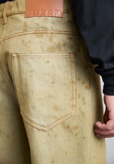 Jeans in denim giallo chiaro con superficie texturizzata, dotati di una toppa in pelle marrone e cuciture arancioni; provvisti di una tasca posteriore.