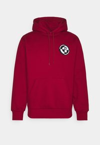 Red hooded sweatshirt made of soft fabric, featuring a front pocket, drawstrings, and a circular embroidered logo on the chest.
