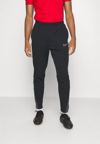 Pantalons de sport noirs avec une taille élastique, une coupe fuselée, des rayures sur les côtés et un petit logo blanc de Nike. Associés à des chaussures de sport noires.
