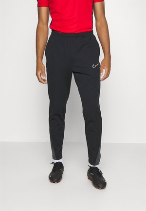 Pantalons de sport noirs avec une taille élastique, une coupe fuselée, des rayures sur les côtés et un petit logo blanc de Nike. Associés à des chaussures de sport noires.