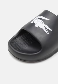 Papuci din cauciuc negru cu vârful deschis, având un logo alb de crocodil, un model texturat pe baza piciorului și margini rotunjite.