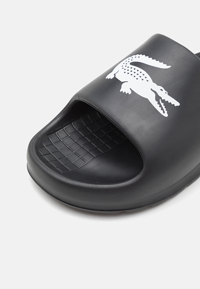 Chausson noir en caoutchouc avec une ouverture à l'avant, arborant un logo crocodile blanc, un motif de grip texturé sur le support de pied et des bords arrondis.