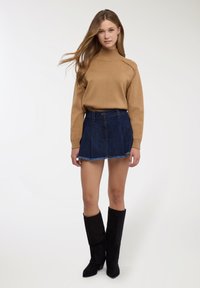 Maglione marrone chiaro lavorato a maglia con collo alto, abbinato a una minigonna in denim scuro con orlo sfrangiato, e stivali alti in suede nero.