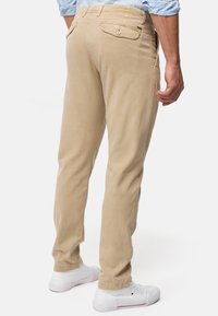 Beige puuvillapantsit, joissa on kapea istuvuus, varustettu takataskuilla, joissa on napilla suljettavat läpät, yhdistetty valkoisiin lenkkareihin. Sileä pinta ja muotoiltu suunnittelu.