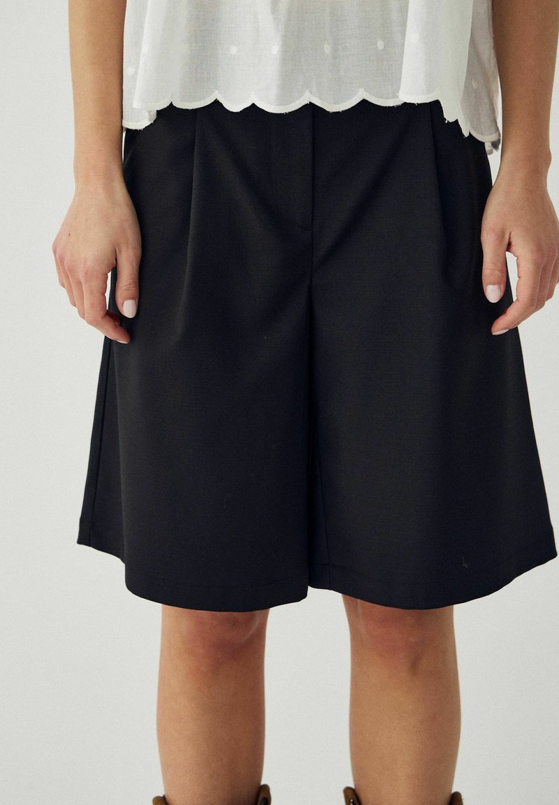 Femme portant un short noir large à mi-cuisses et un haut blanc à ourlet festonné, debout les mains détendues le long du corps.