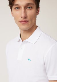 Witte polo met een gestructureerde kraag, twee turquoise knopen en een klein geborduurd turquoise hondenlogo op de borst.