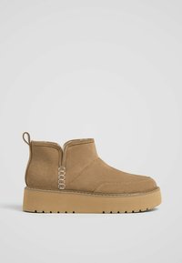 Bottines en suede beige avec une semelle plate, panneaux latéraux élastiques et une languette à boucle. Présentent des accents de couture et une finition texturée.