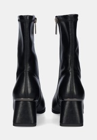 Botas de tobillo de cuero negro con un alto tacón cuadrado, cierre de cremallera y textura suave. Presentan líneas limpias y acentos de hardware mínimos.