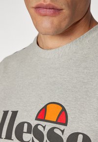 Camiseta de algodón gris con cuello redondo que presenta un gran logo gráfico en negro y naranja. Textura suave y corte relajado.
