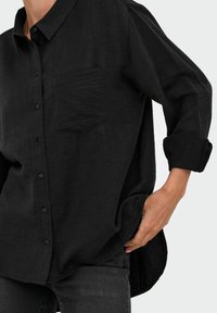 Camisa negra de botones con una tela texturizada, que presenta un bolsillo en el pecho y puños enrollados, combinada con jeans de mezclilla oscura.