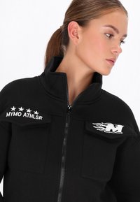 Jachetă neagră cu fermoar, guler înalt, două buzunare la piept, text brodat alb și design cu flacără, confecționată dintr-un material neted și texturat.