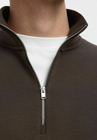 Hnědý pulovr na zip s vysokým límcem, s stříbrným kovovým zipem a bílou vnitřní vrstvou viditelnou na límci. Hladká textura.