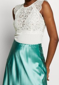 Kvinna i ärmlös vitt spetslinne ihop med högmidjade, glänsande turkosa satängkjol, står mot en plain vit bakgrund.