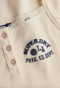 Kremowa dzianina prążkowana z koronkową lamówką, trzema perłowymi guzikami i granatowym haftem z napisem "SUPERDRY LA PHYS. ED. DEPT" oraz ikoną siatkówki.