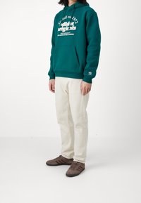 Teal huvtröja med vit grafisk text, beige byxor och bruna sneakers med mörka accenter, uppställd mot en vit bakgrund.