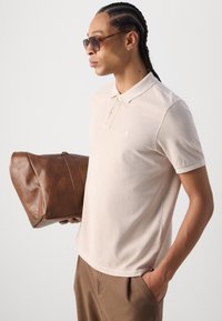 Beige polo-skjorte med korte ermer, knappet krage og liten logo; holder en brun lærdukke, iført mørke solbriller.