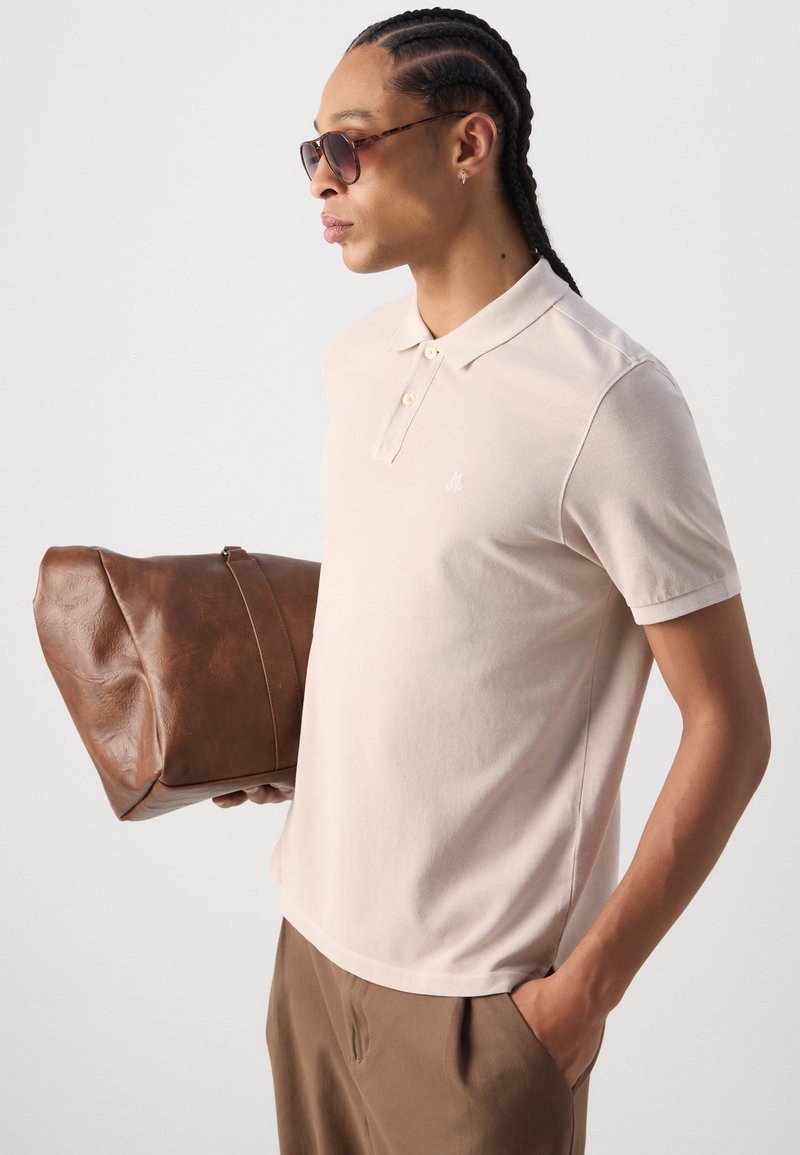 Beżowa koszulka polo z krótkim rękawem, zapięta pod szyją, z małym logo; trzyma brązową skórzaną torbę podróżną, nosi ciemne okulary przeciwsłoneczne.