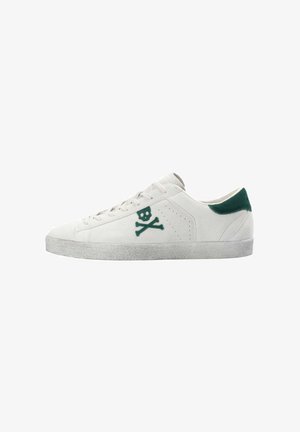 Zapatilla de ante blanca con un tabique de talón de ante verde, que presenta un logo de calavera y huesos cruzados verde en el costado y una suela de goma texturizada gris.