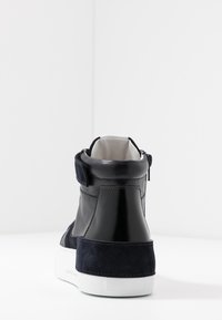 Laivastonsininen nahka- ja mokkanahkainen korkeavartinen lenkkari, jossa on valkoinen kumipohja. Kenkä on sileä pinta, poikkeavat tekstuurit ja takana vetoketjukiinnitys.