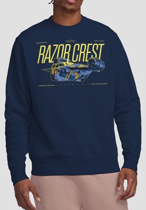 Navy sweatshirt with yellow text "RAZOR CREST" and illustration of a spaceship from Mandalorian, worn by a person with dark skin and pink pants.