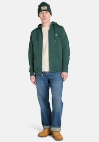 Felpa verde con zip e logo frontale, camicia crema sotto, jeans blu e stivali color beige. Presenta due tasche frontali e cordini regolabili.