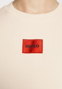 Gros plan d'une chemise beige avec un patch rectangulaire rouge embossé du mot « HUGO » en lettres noires sur la poitrine.