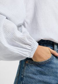 Blusa a righe blu chiaro con maniche a palloncino e polsini elastici, abbinata a jeans in denim con tasca frontale e cuciture a vista.
