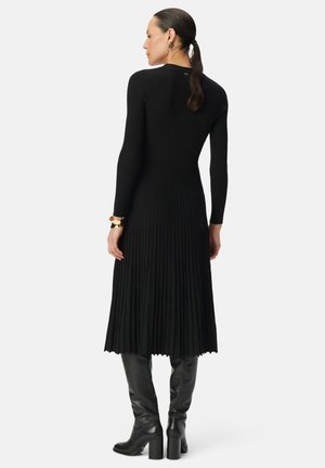 Schwarzes, geripptes Strickkleid mit langen Ärmeln und einem ausgestellten, plissierten Rock. Mit einem offenen Schlüsselhole auf der Rückseite und getragen mit kniehohen schwarzen Stiefeln.