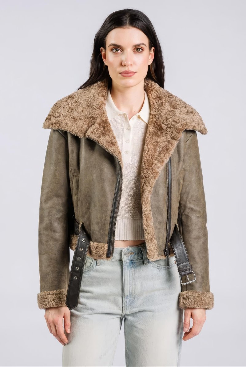 Vrouw met lang donker haar draagt een bruine shearling jas over een crèmekleurige gebreide top en lichtgewassen jeans tegen een effen grijze achtergrond.