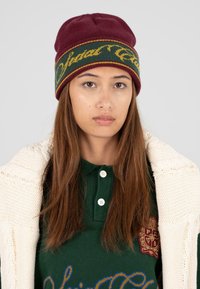 Bordeaux gebreide beanie met groene gestreepte band met gele tekst. Gedragen met een donkergroen polo-shirt en een gestructureerde crèmekleurige cardigan.