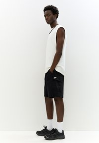 Hvid ærmeløs top, sorte cargo shorts med lommer, sorte sneakers og hvide crew-sokker. Modellen står foran en lys baggrund.