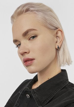 Pendiente de botón en tono plateado con un intrincado diseño floral, llevado en una oreja. La modelo tiene el cabello rubio claro y viste una chaqueta de mezclilla negra. Su tono de piel es claro.