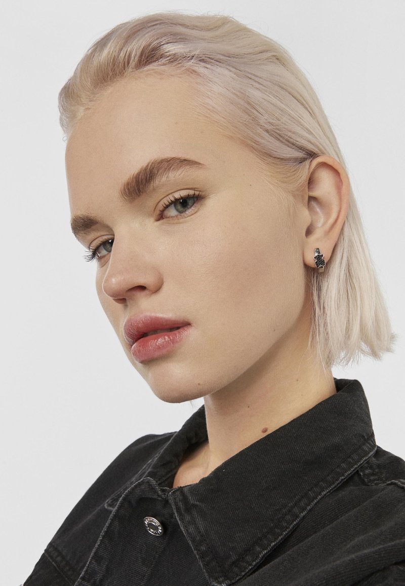 Pendiente de botón en tono plateado con un intrincado diseño floral, llevado en una oreja. La modelo tiene el cabello rubio claro y viste una chaqueta de mezclilla negra. Su tono de piel es claro.