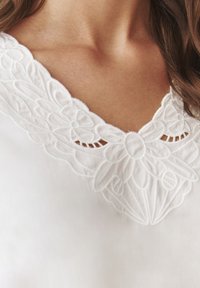 Blusa bianca con intricati ricami floreali sul collo, con bordo scallop e texture morbida, che esalta la semplicità del tessuto.