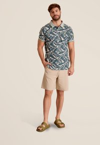 Korte mouw polo met een groen en crème bladerenpatroon, contrasterende kraag met gele strepen, gecombineerd met beige shorts en groene sandalen.