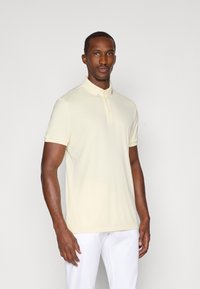Beige polo shirt med krave, korte ærmer og treknaps lukning, lavet af glat stof; båret med hvide bukser; ingen mønstre.