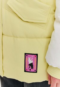 Nafukovacia, žebrovaná bunda v svetložltej farbe s čiernym štvorcovým nášivom s dizajnom mačky a farebným textom. Hladká textura s vrstveným dizajnom.
