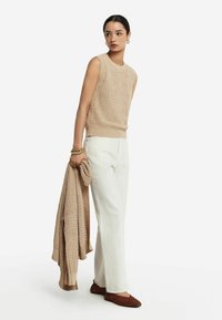 Beige strikket trøje med blomster mønster, parret med hvide bukser og brune flade sko. Modellen holder en matchende trøje i den ene hånd.