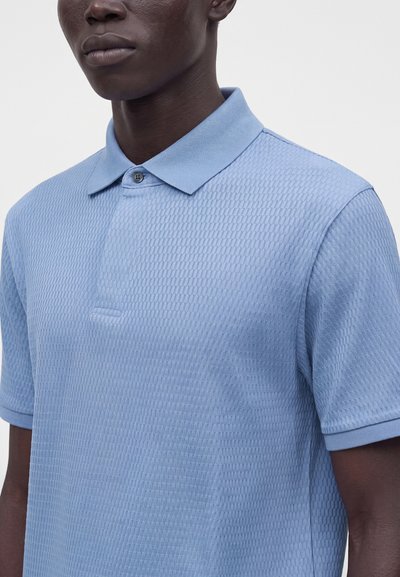 Homme portant un polo bleu clair texturé à manches courtes, avec un col et un seul bouton au niveau du cou.
