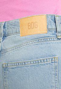 Vaqueros de mezclilla azul claro con un parche de cuero etiquetado "BDG" en la cintura, que muestra una textura desgastada y costuras en contraste.