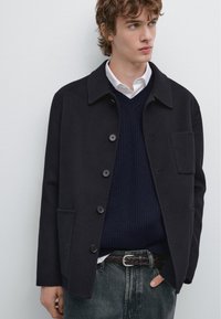 Sort uldblandingsjakke med krave, knaplukning og sidelommer, iført over en marineblå striktrøje og mørke jeans.