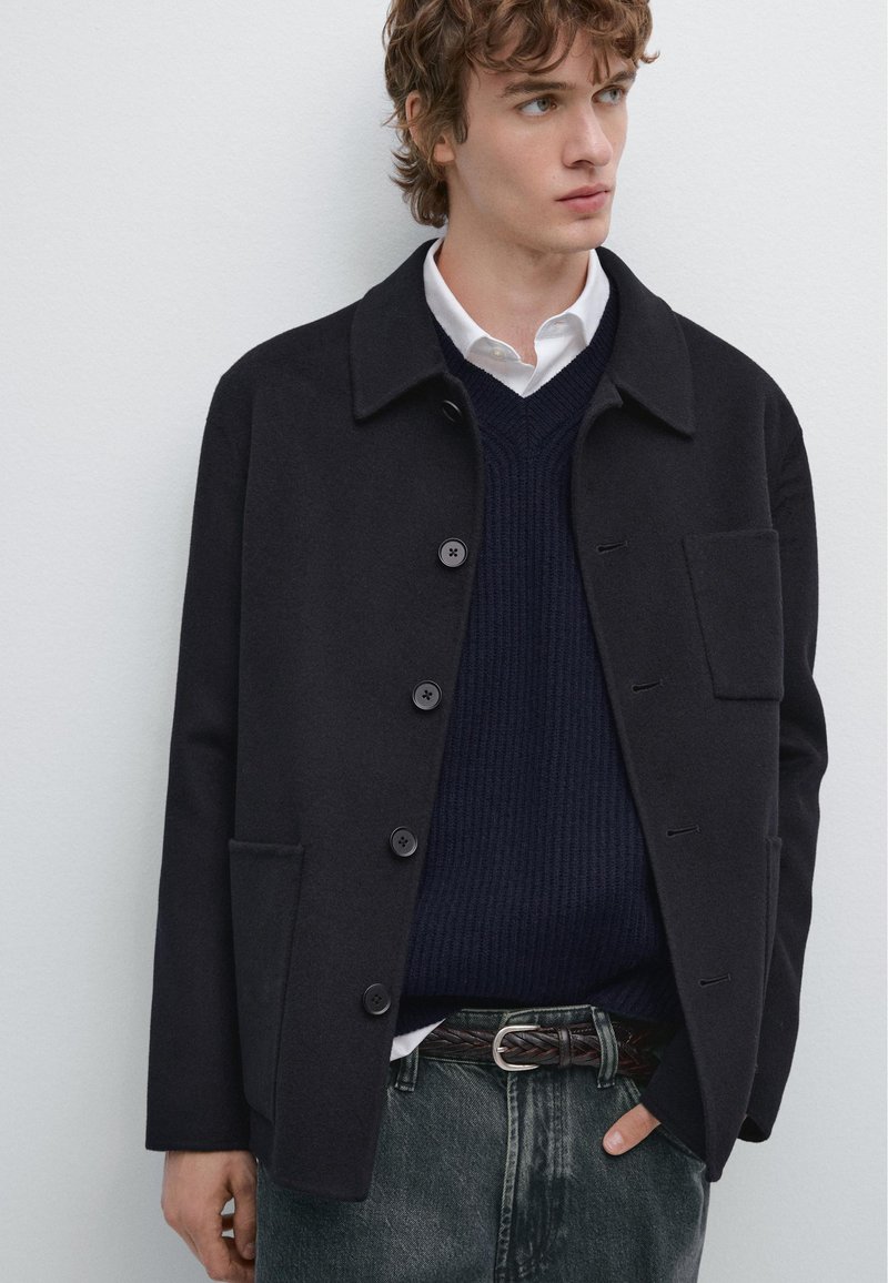 Sort uldblandingsjakke med krave, knaplukning og sidelommer, iført over en marineblå striktrøje og mørke jeans.