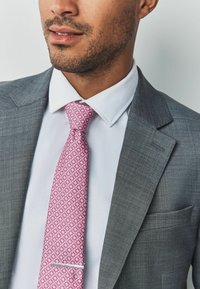 Grau Anzug mit einem hellen Hemd und einer rosa gemusterten Krawatte. Die Krawatte zeigt florale Designs; der Anzug hat einen strukturierten Stoff und eine Tasche.