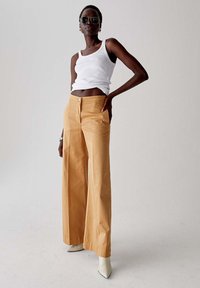 Pantaloni a gamba larga color senape con chiusura a un bottone, abbinati a un top aderente bianco. Modella con occhiali da sole e tacchi beige.
