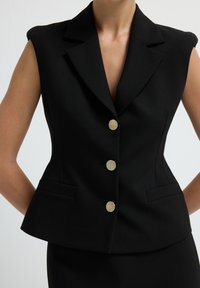 Veste sans manches noire sur mesure avec un col structuré, des boutons dorés et deux poches latérales, présentant une silhouette ajustée et une texture lisse.