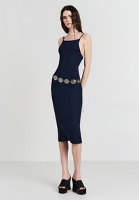 Femme portant une robe midi bleu marine à fines bretelles, une ceinture en pièces argentées et des sandales à plateforme noires, se tenant sur un fond blanc.