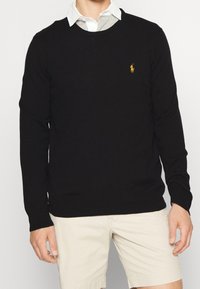 Man i en svart tröja med rund hals över en vit skjorta med krage och beige shorts, som står framför en enfärgad bakgrund.