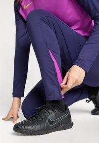 Tenue de sport violette et marine, comprenant un legging avec des accents roses sur les côtés. La personne ajuste le poignet du pantalon près d'une chaussure de soccer noire.