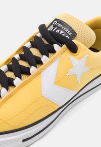 Baskets en toile jaunes avec lacets noirs, logo étoile blanche, languette rembourrée et semelle extérieure en caoutchouc noir, avec des accents de couture blanche.